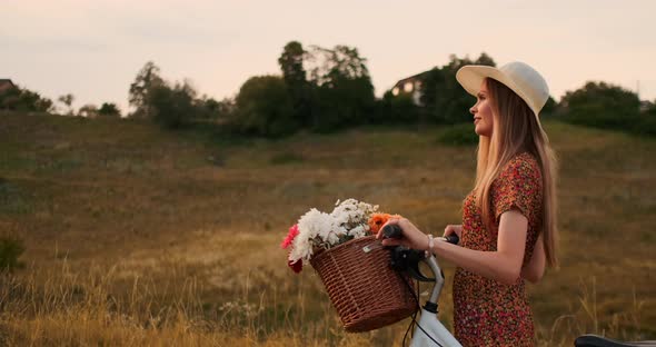 Young Smiling Blonde in Hat and Dress Walking in Dress with Bike and Flowers in Basket