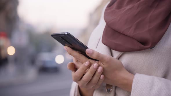 A Muslim Woman Works Quickly on a Smartphone in a Street in an Urban Area  a Road in the Background