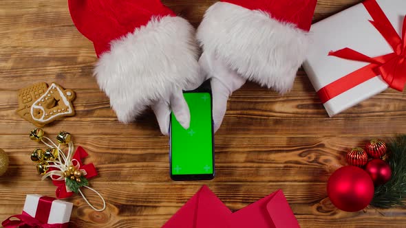 Top View Santa Hands Uses Smartphone with Green Screen Chroma Key By Wooden New Year Decorated Table