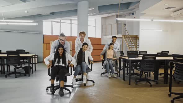 Young Group of Diverse Colleagues are Celebrating Success Racing on Chairs in Office Laughing and