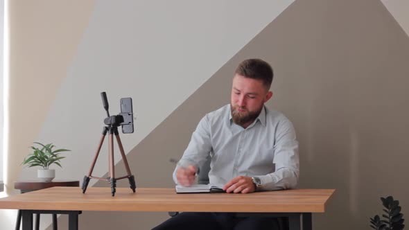 Man with a Beard in a Blue Shirt Sits at His Desk in Room Blogs on Phone Online