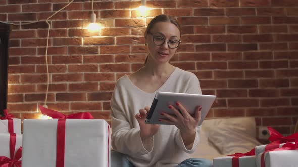 Positive Woman Glasses Checks Gifts Using Smart Tablet Then Looks Camera