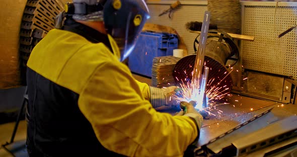 Welding Works at the Plant. Employee in a Protective Mask, Suit and Gloves Welds Metal Parts with