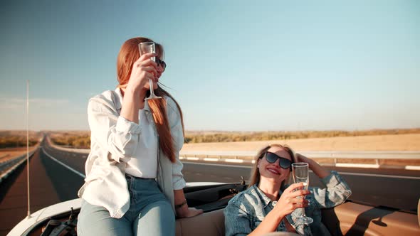 Two joyful female students are driving car without roof on vacation to beach and drinking beverages.