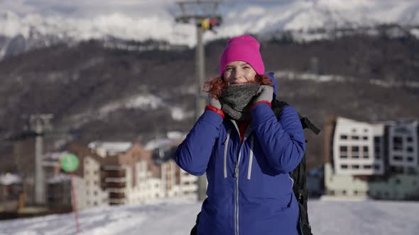 A Woman in a Pink Hat and Blue Jacket Stands and Smiles on a City Winter Street Against the Backdrop