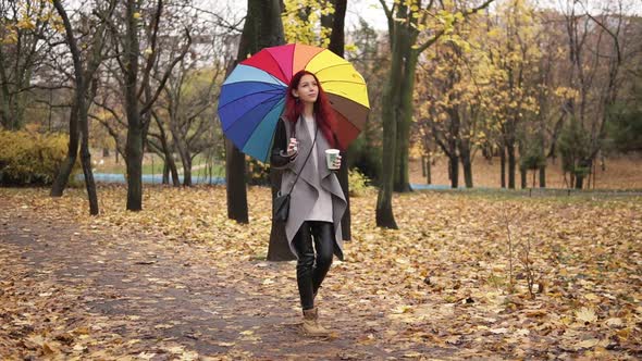 Young Stylish Woman with Red Hair Walking in Autumn Park and Drinking Coffee From a Paper Cup While