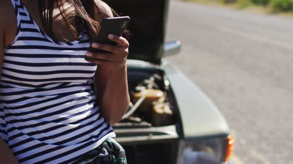 African american woman using smartphone while standing near her broken down car on road