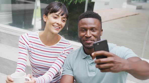 Happy diverse couple wearing blouse and shirt and taking selfie with smartphone in garden