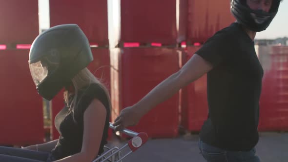 Guy Rolls Attractive Young Woman in the Trolley in Motorcycle Helmets at Sunset