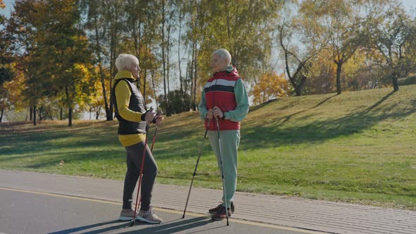 Two Aged Women Talk in Sunny Fall Park Wearing Vests and Holding Walking Poles