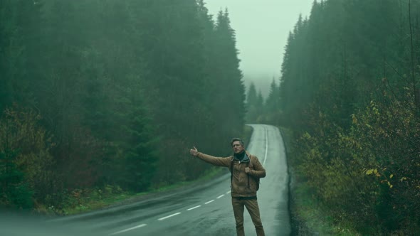 Man Hitchhiker Traveler with Backpack Stands at Mountain Road Outdoors at Forest in Cold Wet Foggy