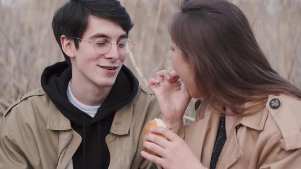 Girl in Coat Feeds Boyfriend Croissant on Picnic in Field