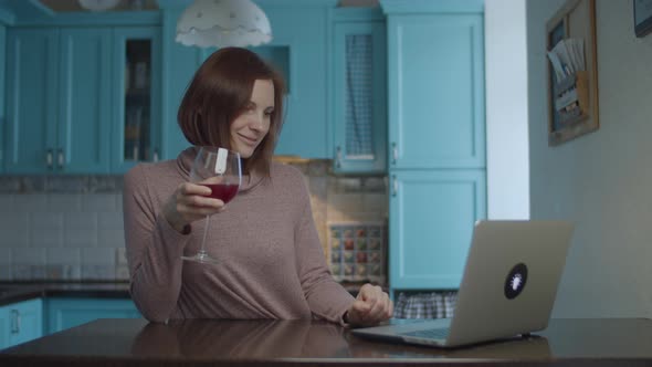 Young 30s smiling woman drinking glass of red wine and typing on laptop
