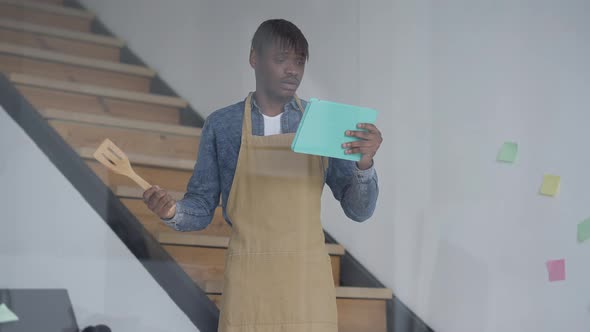 Young African American Man in Apron Gesturing with Cooking Shovel Talking at Tablet Web Camera