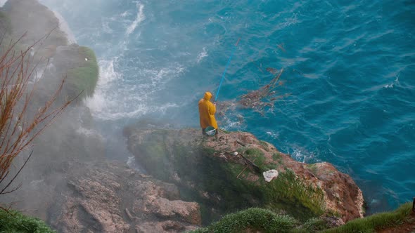 A Man in a Yellow Raincoat Fishing Standing on the Stones By the Waterfall