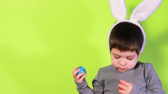 A Boy with Bunny Ears on His Head in a Gray Jacket Plays with Colorful Eggs