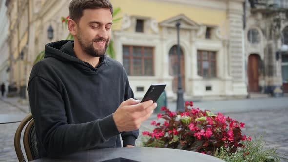 Hipster Guy with Mobile Smart Phone Digital Camera Talks While Sitting in Cafe