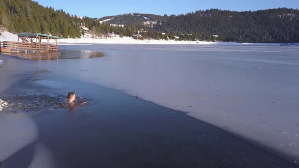 Aerial views of man sitting neck deep in frozen mountain lake, drone rotating. Young guy doing polar