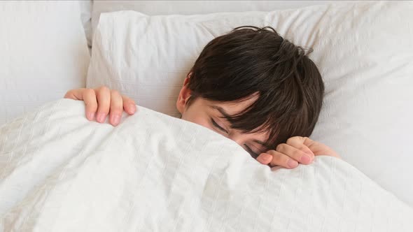 Young woman with short hair peeks out from under the covers waking up in bed in the morning.