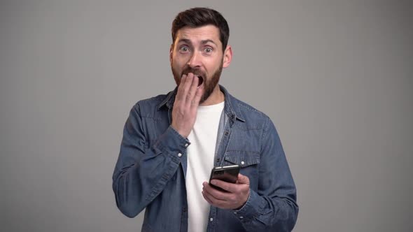 Portrait bearded man in shirt looking at smartphone display with widely open mouth, shocked