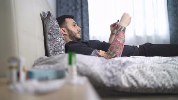 Hipster with Tatoo Using Thermometer Lying in Bed