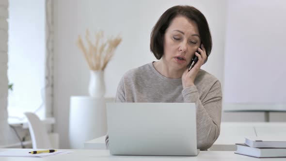 Old Senior Woman Talking on Phone and Working on Laptop