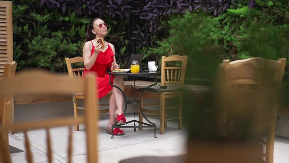 Wide Shot of Happy Beautiful Young Caucasian Woman in Red Dress and Sunglasses Eating Burger