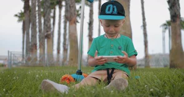 Young Boy Watches a Video on His Mobile Phone Sitting on the Grass in Park, Children and Modern