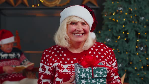Senior Grandmother in Festive Sweater Presenting Christmas Gift Box Smiling Looking at Camera