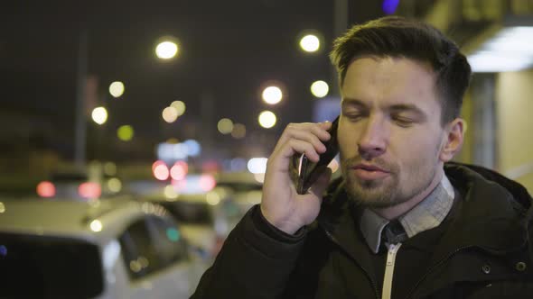 A Young Handsome Man Talks on a Smartphone with a Smile in a Street in an Urban Area at Night