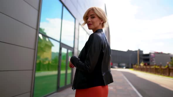 the Blonde Goes Along the Wall with Large Windows, Turns Around and Takes Off Her Black Jacket