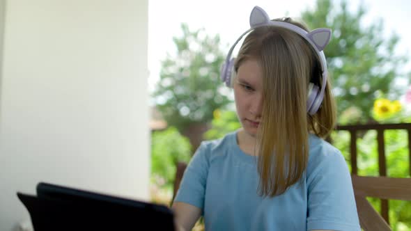 Teenage Girl Sits At Wooden Table In a Summer Cafe In Headphones With Phone