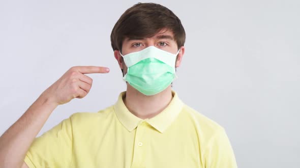 Serious Man in Yellow Shirt and in Protective Respiratory Mask Pointing on His Face Isolated on Grey