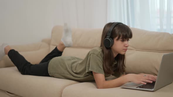 A Teen Girl Uses a Laptop While Lying on a Couch in Headphones