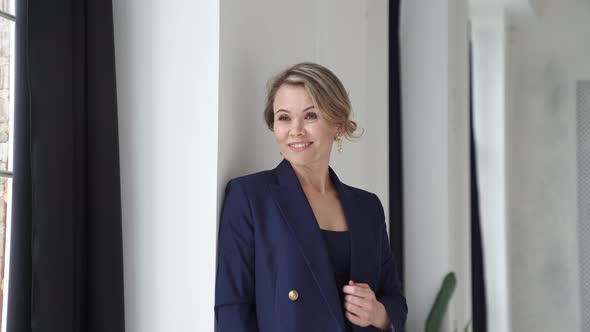 A Blonde Woman in a Blue Suit Poses Standing By a Window in a Photo Studio