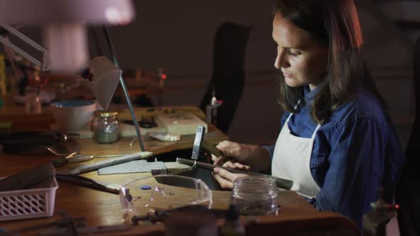 Profile of focused caucasian female jeweller sitting at desk, making jewelry in workshop