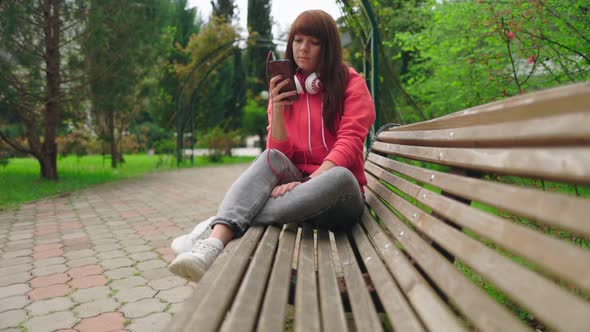 A young woman with headphones, sitting on a bench in the park and using the phone in her hands.