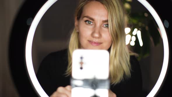 Adorable blonde woman streaming beauty vlog on mobile phone with ring lamp from home