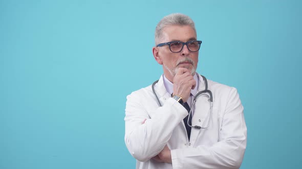 Elderly Man Doctor in Glasses Thoughtfully Rubs Chin with Hand Standing in the Studio on a Blue