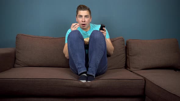 A Young Man Is Watching TV and Eating Popcorn. The Guy Is Watching TV Carefully. An Exciting Moment