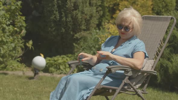 Carefree Female Retiree Sitting in Garden Chair and Applying Hand Moisturizer. Portrait of Happy