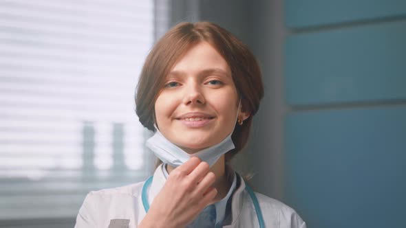Brunette young woman doctor takes off blue disposable mask and looks straight smiling
