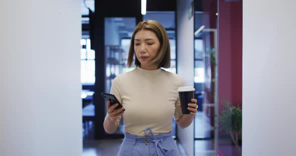 Asian businesswoman walking using smartphone holding cup of coffee in modern office