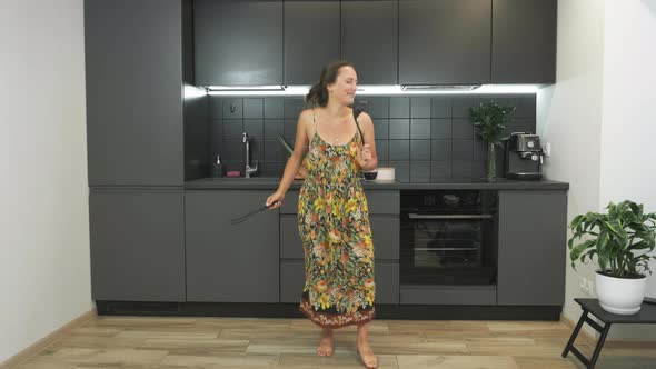 Cute joyful caucasian young woman in home wear is having fun and dancing with kitchenware