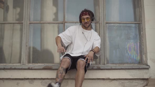 Confident Male Break Dancer with Tattoos and Dreadlocks Sitting on Urban Windowsill in Headphones