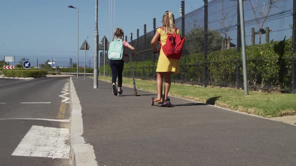 Two girls with school bags riding scooters on footpath