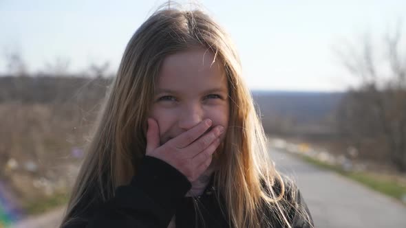 Close Up of Happy Little Girl Looks Into Camera Outdoor and Laughs Covering Her Face with Hand