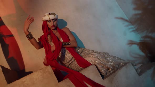 a Blonde in a Turban and a Shiny Dress Sits on the Stairs and Moves Hands Beautifully with a Red