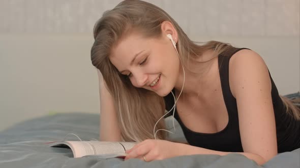 Cute Female with Headphones Reading a Magazine While Lying on a Bed