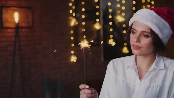 Pretty Woman in Santa Claus Hat and Shirt Flirts at Camera with Sparklers
